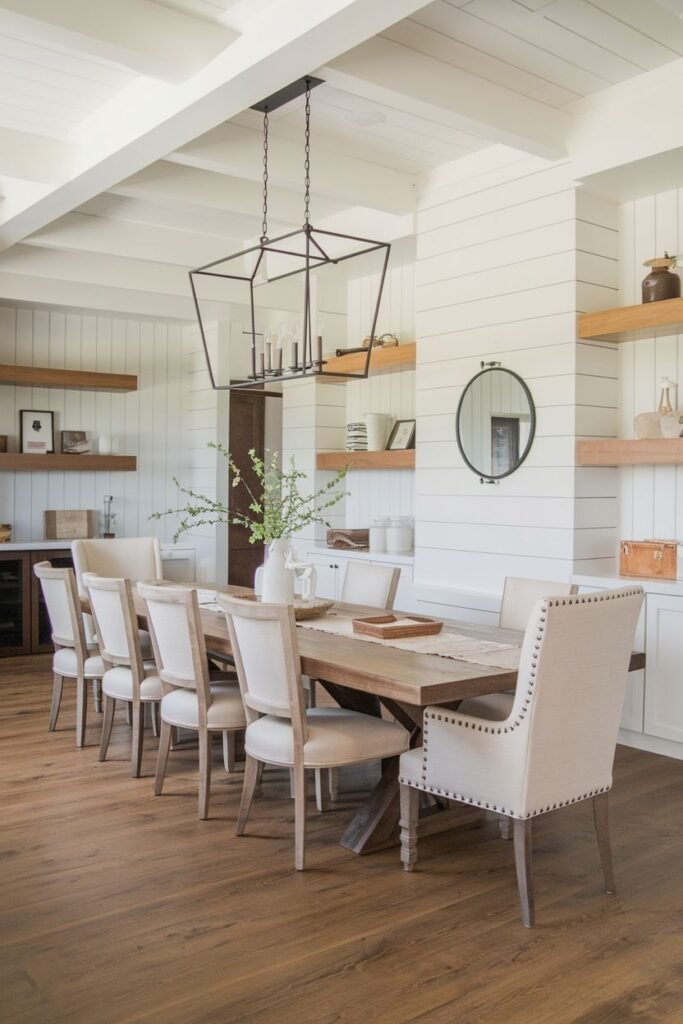 A beautiful farmhouse dining room with a rectangular wood table and upholstered chairs with nailhead trim. The room features white shiplap walls, exposed beams, open shelving, and a large black rectangular chandelier.