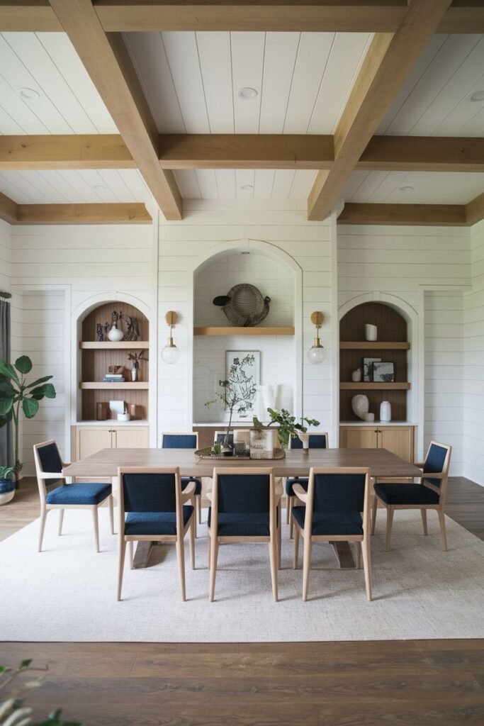 A beautiful farmhouse dining room featuring a light wood rectangular table surrounded by chairs with blue upholstered seats. The room has white shiplap walls, exposed beams, and built-in arched shelving units.