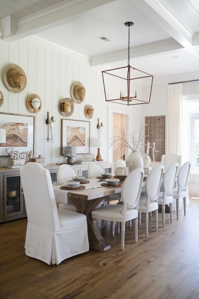 A beautiful farmhouse dining room featuring a light wood rectangular table with a grey wood base and multiple white slipcovered chairs, set on wood floors. The room has white shiplap walls, wall art, and several straw hats hung as decoration.