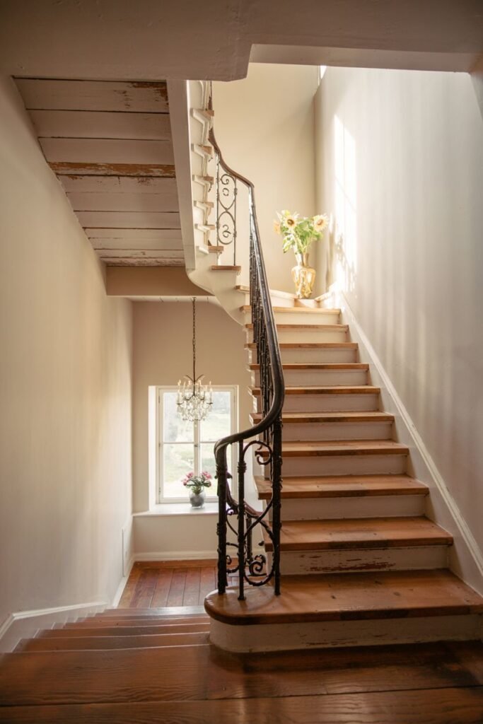 Charming curved stair with white risers and dark wooden treads, featuring an ornate dark metal railing and sunlight streaming through a window.