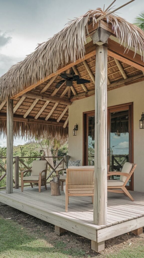 A rustic wooden deck covered by a roof thatched with straw or palm leaves, supported by round wooden posts. The deck is furnished with comfortable outdoor chairs and a coffee table. A wooden railing surrounds the deck. The background shows greenery and hills.