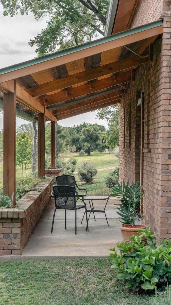 A concrete patio next to a brick house. The patio is covered by a wooden roof with exposed beams. Simple metal chairs and a small table are on the patio, along with potted plants. Low brick walls form a border for planting beds.