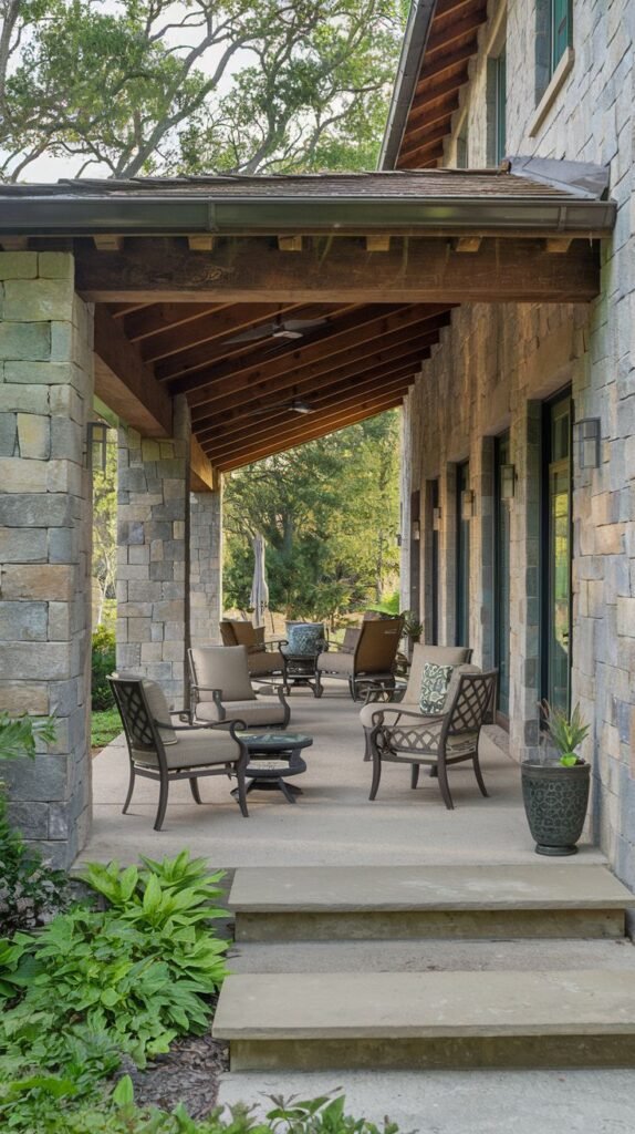 A covered patio constructed with large stone pillars and walls. The patio features a seating area with comfortable chairs and small tables. Steps lead down from the patio. Beyond the seating area, more chairs are visible, and the background shows trees and greenery.