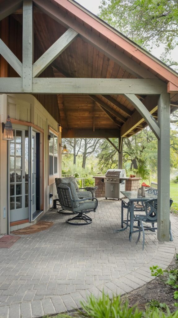 A covered outdoor space with a high wooden roof structure supported by a wooden post. The flooring is brick laid in a circular pattern. There is a seating area with a rocking chair and a separate dining area with a table and chairs. An outdoor grill is also present.