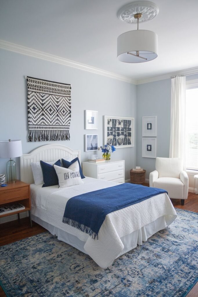 A bedroom with light blue walls, a white arched headboard bed with a navy blue throw and pillows, a white dresser, a cream accent chair, and a patterned woven wall hanging, featuring a large blue and white patterned rug.