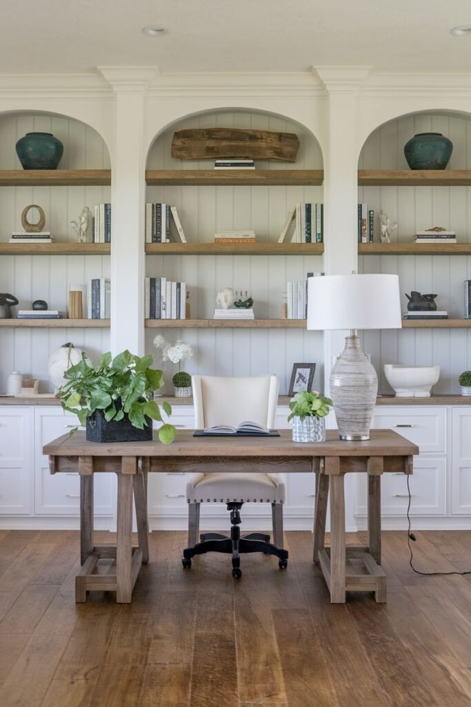 Charming and cozy farmhouse office with white built-in shelves featuring arched openings and a rustic wooden desk with trestle legs.