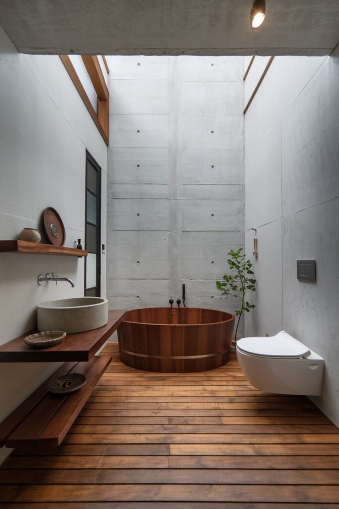 A cozy japandi bathroom with exposed concrete walls and ceiling, a traditional deep wooden soaking tub, wide wooden floor planks, floating wooden shelves with a stone basin sink, and a wall-mounted toilet.