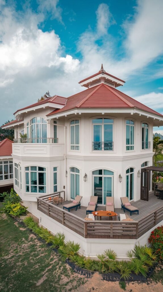 An aerial view of a dreamy luxury white house with a distinctive red tiled roof, featuring bay windows, balconies, a wooden deck with lounge chairs and a table, and surrounding greenery, under a cloudy blue sky.