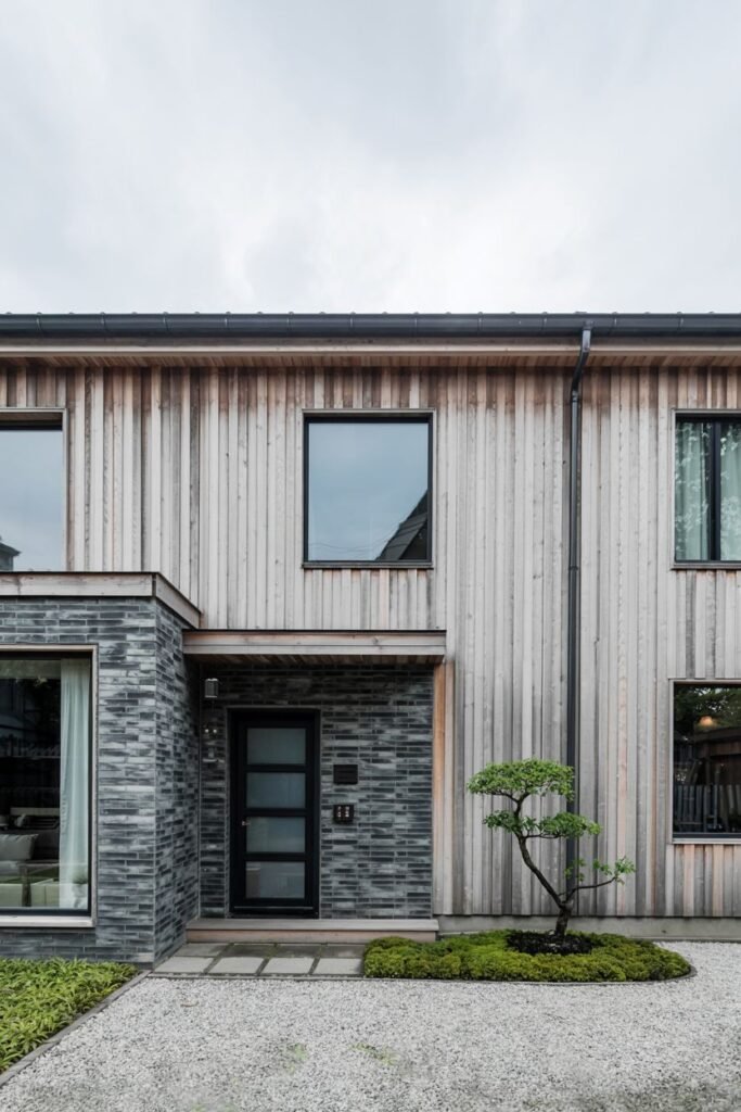 Mixed materials on a Contemporary japandi house exterior with vertical wood siding, dark brick/stone cladding, a dark door with glass panels, and a gravel entrance with a moss garden.