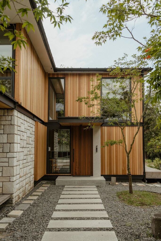 Contemporary japandi house exterior showing a contrast of light stone blocks and extensive warm wood siding, with a pathway of stepping stones over gravel and rocks.