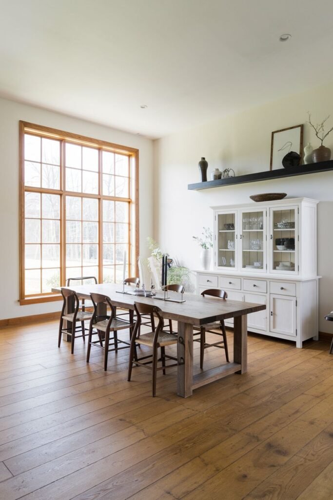 A dining room with a wooden table and eight wooden chairs has a large white china cabinet and a large multi-pane window.