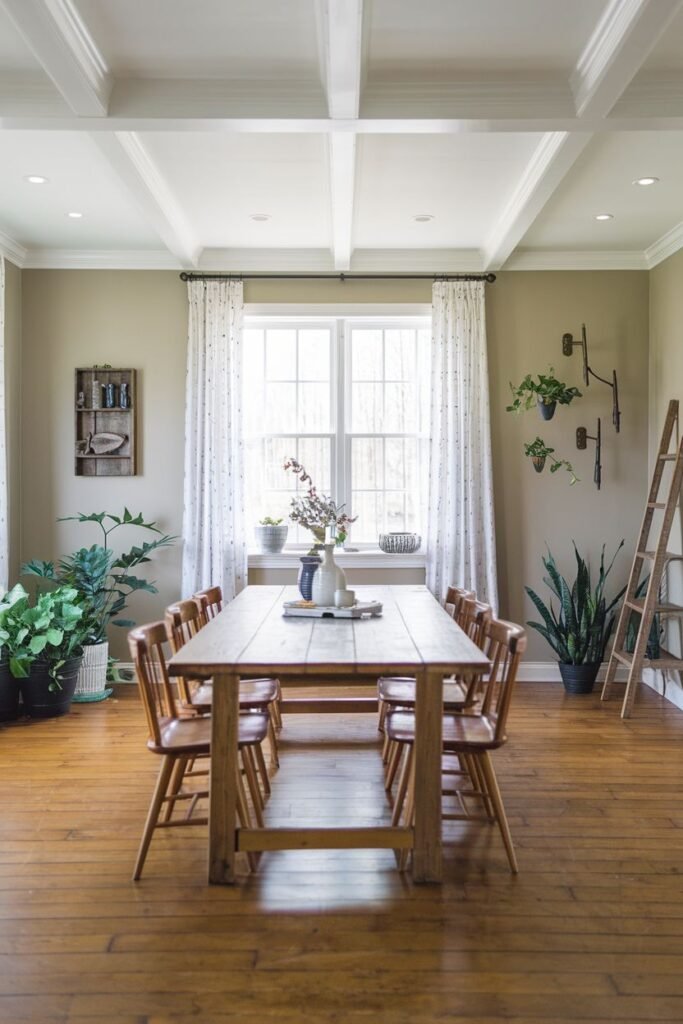 A dining room features a light-colored wooden table with eight wooden chairs, a coffered ceiling, plants, a decorative ladder, and wall art.