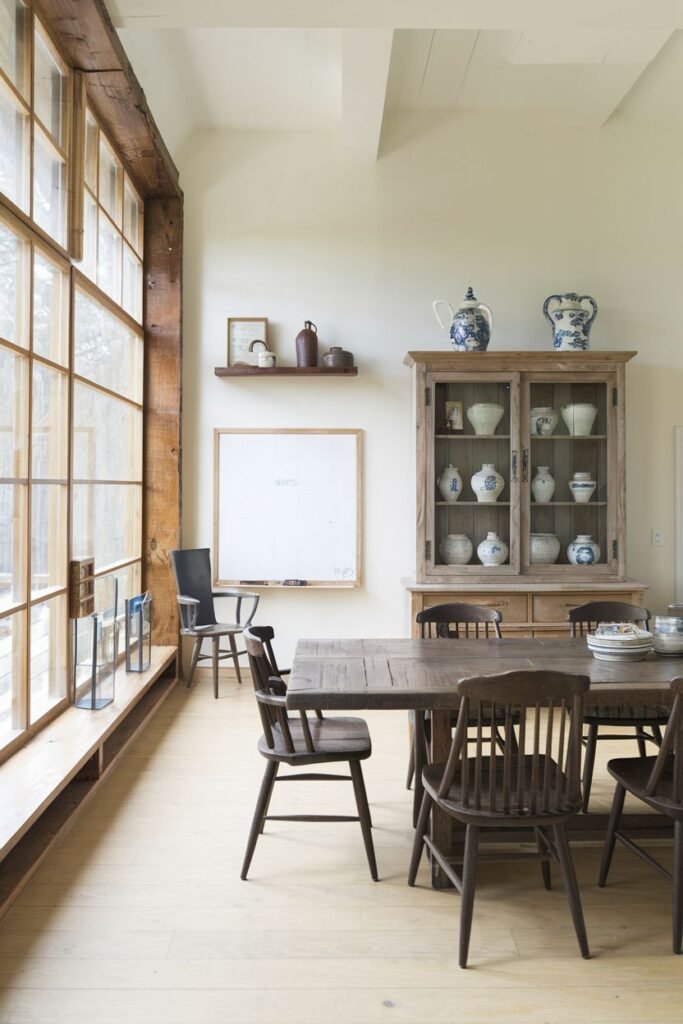 A dining area has a dark wooden table and five chairs near a large window, with a wooden cabinet filled with pottery and a whiteboard.