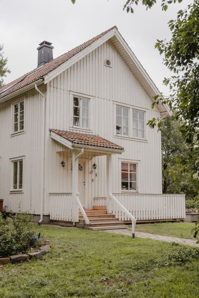 A traditional white wooden Scandinavian house exterior with a covered front entrance, railing, and steps, featuring multiple windows and a classic tiled roof.