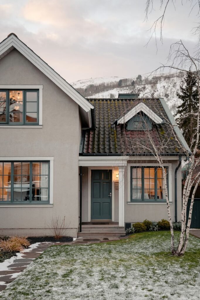 A light grey stucco Scandinavian house exterior with distinctive grey-green window frames and a covered entryway leading to a dark door, set in a partially snow-covered yard with a mountain backdrop.