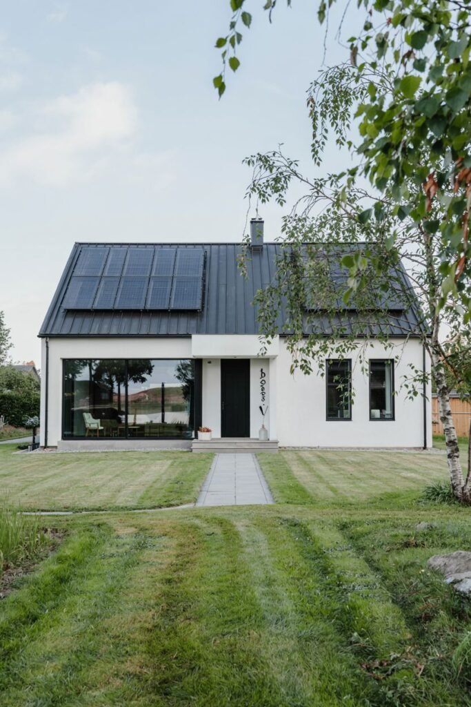 A minimalist white Scandinavian house exterior with a bold dark roof covered in solar panels and a large corner window, with a straight walkway leading to the front door.