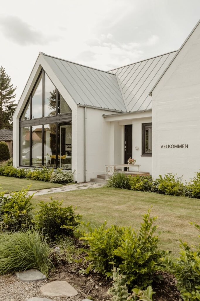 A clean white Scandinavian house exterior with a modern angled roofline and a large angled window that follows the roof shape, with a bench near the entrance and a "Velkommen" sign on the wall.