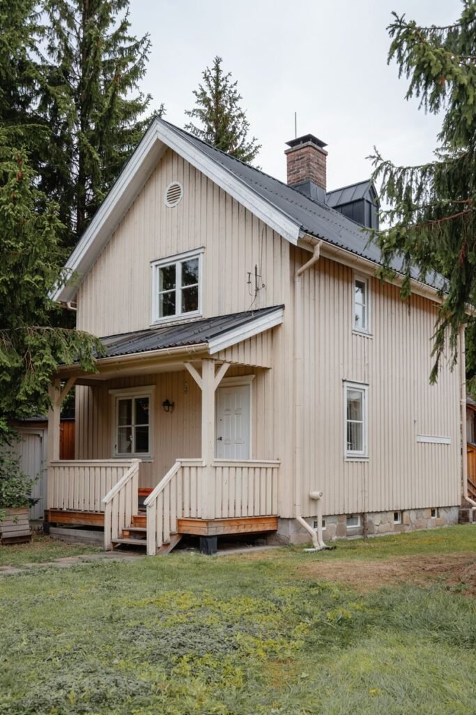 A pale yellow vertical-sided two-story Scandinavian house exterior with a covered front porch and grey pitched roof, surrounded by trees.