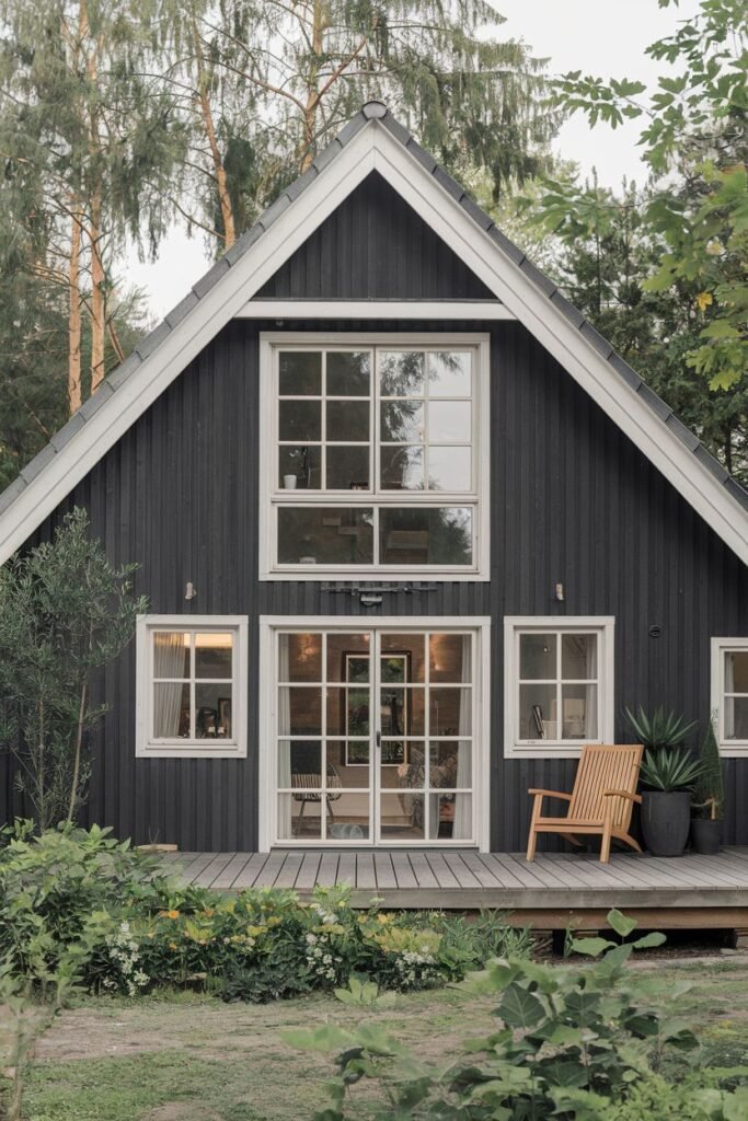 A dramatic dark-sided Scandinavian house exterior with white window frames and trim, featuring a large wooden deck with outdoor seating and planters accessible through expansive glass doors.