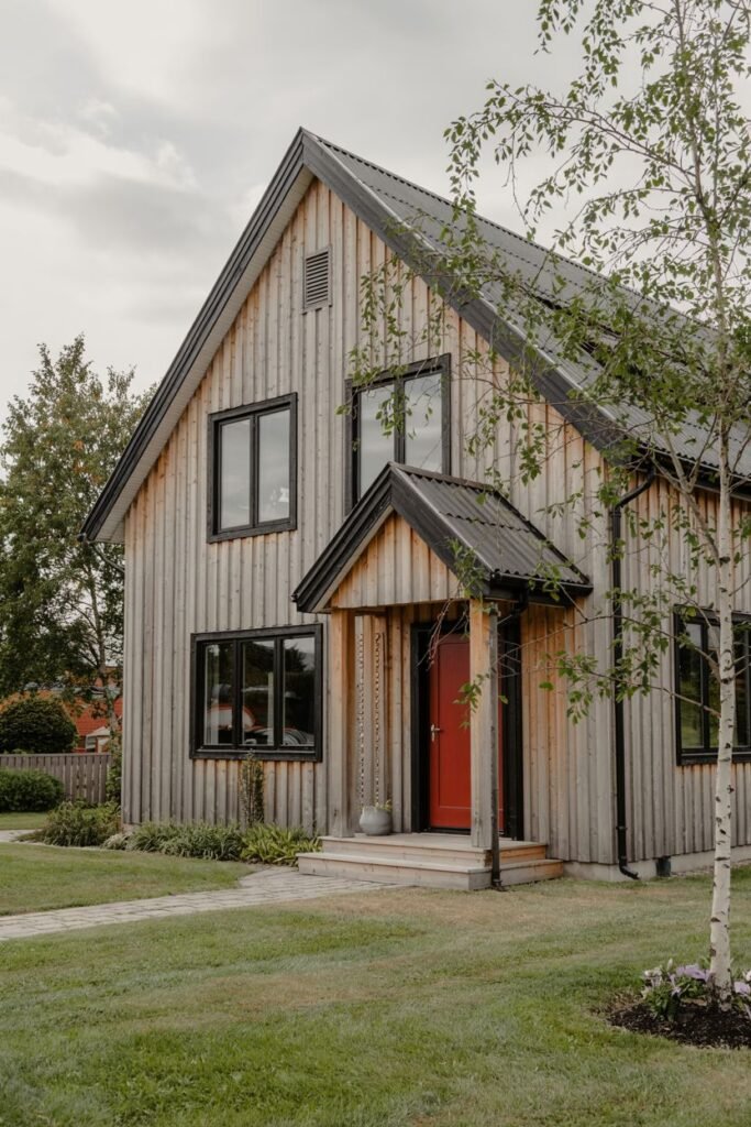A natural wood-sided Scandinavian house exterior with a dark roof, black window frames, and a bright red front door under a small covered entryway, with a tree in the foreground.