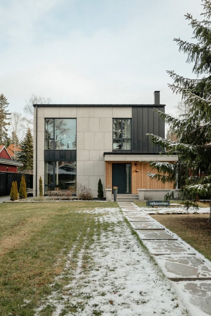 A modern Scandinavian house exterior combining light panels and dark metal siding with wood cladding around the entrance, featuring large windows and a path with a "Velkommen" sign, set in a wintery landscape.