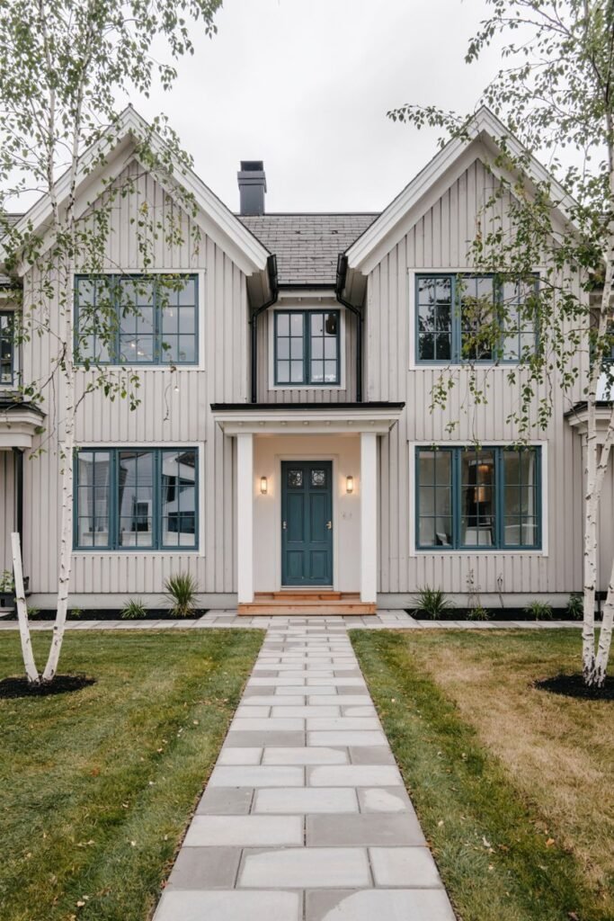 An elegant light grey vertical-sided Scandinavian house exterior with dark teal double front doors, symmetrical window placement, and a paved pathway leading directly to the entrance.
