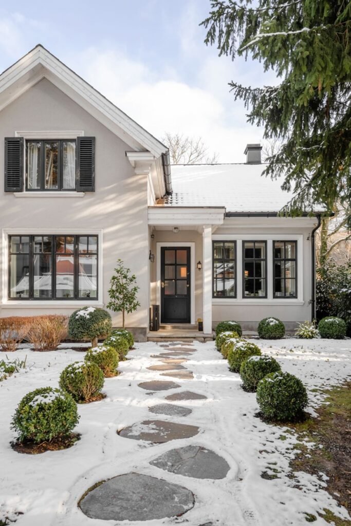 A light stucco Scandinavian house exterior with dark window shutters on the upper level and a path of irregular stepping stones bordered by spherical bushes, set in a snowy yard.