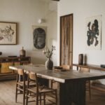 An elegant dining room with a large wooden table and minimalist wooden and woven-seat chairs, featuring artwork and simple furniture, showcasing a wabi sabi aesthetic.